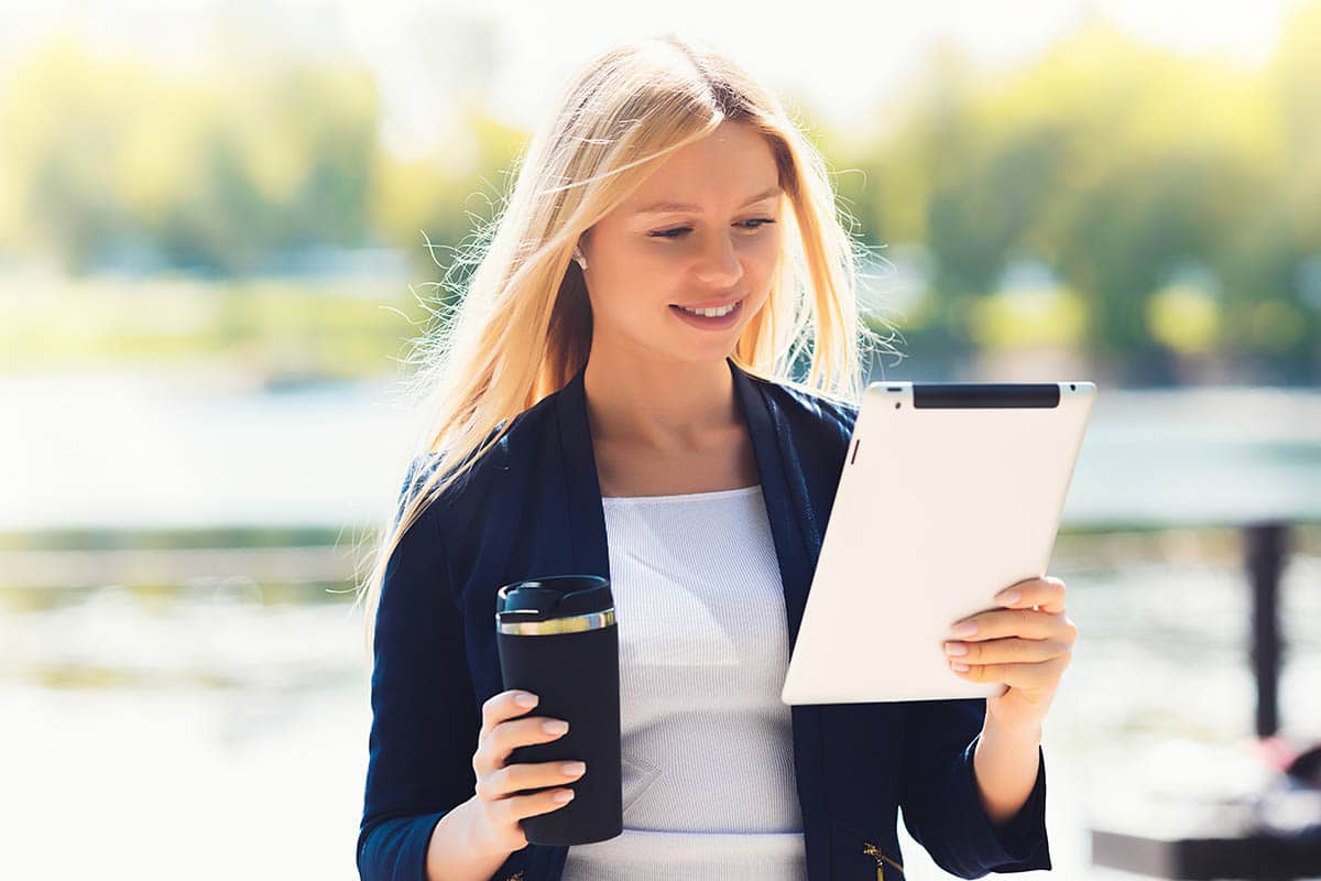 Young women outside with travel mug reading tablet